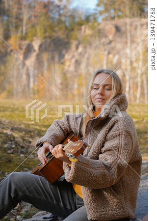 Beautiful blonde girl playing acoustic guitar and singing relaxing enjoying outdoors on sunny autumn day. Hippie woman in boho style learning and practicing playing musical instrument. Fall leaves Beautiful blonde girl playing acoustic guitar and singing relaxing enjoying outdoors on sunny autumn day. Hippie woman in boho style learning and practicing playing musical instrument. Fall leaves 139471788