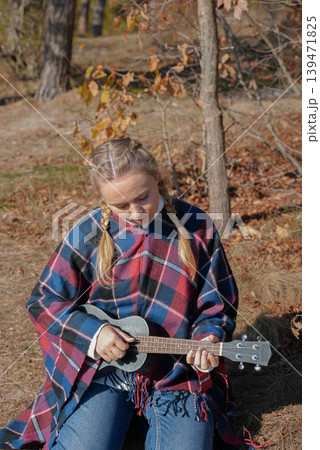 Portrait of smiling caucasian girl plays ukulele in autumnal park. Young woman plays guitar musical instrument outside in nature fall time. Audio music healing Portrait of smiling caucasian girl plays ukulele in autumnal park. Young woman plays guitar musical instrument outside in nature fall time. Audio music healing 139471825