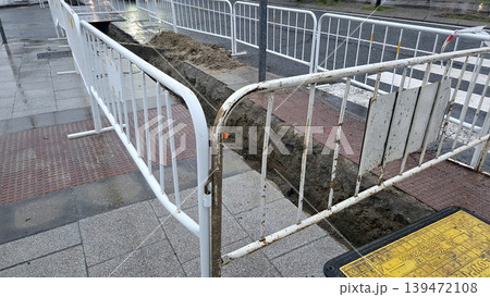 Corner sidewalk trench with metal barriers for pipe laying and road repair at pedestrian crossing for construction maintenance documentation and safety background Corner sidewalk trench with metal barriers for pipe laying and road repair at pedestrian crossing for construction maintenance documentation and safety background 139472108