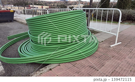 Coiled green plastic conduit tubing stacked on pavement beside a safety barrier for utility installation and pipe laying supply and maintenance work. Telecommunication pipes 139472115