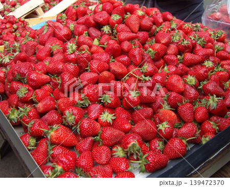 Large, abundant heap of fresh, ripe strawberries fills a market display tray. The vibrant red berries, many with green leafy hulls still attached, present a juicy and natural texture 139472370