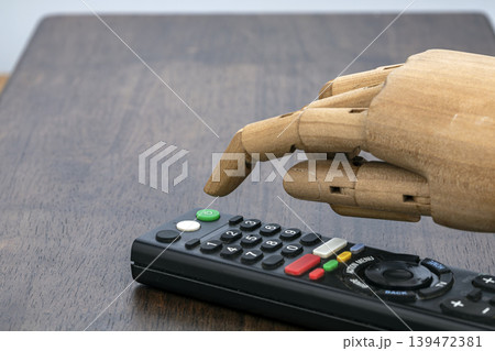 Jointed Wooden Artist Mannequin Hand Pointing Index Finger to Press Button on Black Smart Television Remote Control Resting on Dark Wooden Table Surface Jointed Wooden Artist Mannequin Hand Pointing Index Finger to Press Button on Black Smart Television Remote Control Resting on Dark Wooden Table Surface 139472381