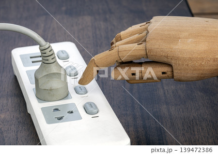 Close-up of an articulated wooden mannequin robot hand with index finger pressing a switch button on a white multi-outlet power strip surge protector on dark wood Close-up of an articulated wooden mannequin robot hand with index finger pressing a switch button on a white multi-outlet power strip surge protector on dark wood 139472386
