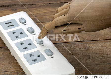 Close-up of a detailed wooden articulated robot hand with its index finger pressing one of the individual switches on a modern white multi-outlet power strip placed on rustic wooden surface 139472388