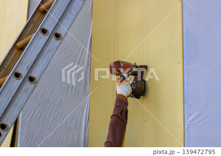 Worker is using nail gun to fasten siding panels on an exterior wall while positioned ladder at construction site. Worker is using nail gun to fasten siding panels on an exterior wall while positioned ladder at construction site. 139472795