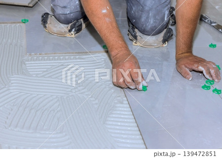 Worker places tiles on floor using cement while adjusting spacers for alignment in home renovation project. 139472851