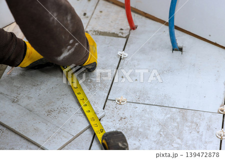 Worker uses measuring tape to check tile placement on floor while doing construction work in new house Worker uses measuring tape to check tile placement on floor while doing construction work in new house 139472878