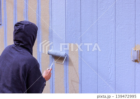 Worker is rolling blue paint on wooden siding wall in home setting during works day. 139472995
