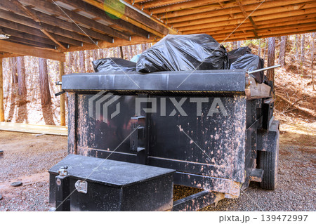 Black dumpster is located under wooden shelter it is filled with garbage bags Black dumpster is located under wooden shelter it is filled with garbage bags 139472997