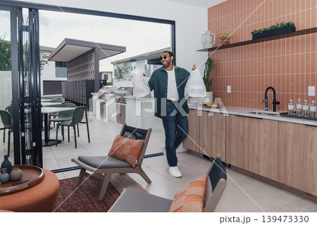 Man Entering Sunlit Open Kitchen Living Space, Carrying Coffee And Wearing Sunglasses With Blazer, Floortoceiling Glass Doors Opening To Patio, Terracotta Tiles And Wooden Cabinets, Leather 139473330