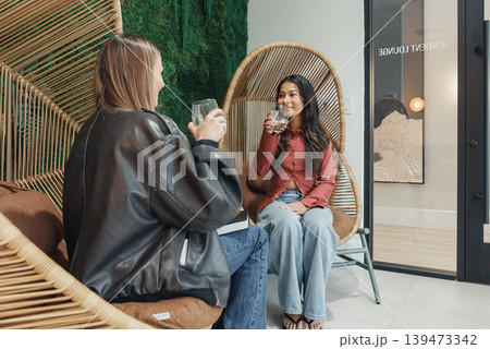 White And Asian Women Clinking Glasses For Brand Shoot, Content Creators Posing In Rattan Chairs With Editorial Styling, Leather Jacket And Blouse Outfits, Studio Backdrop With Poster, 139473342