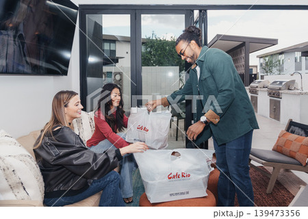 Delivery Driver Handing Takeout Bags To Two Friends On Sofa During Casual Home Meetup, Branded Bags On Ottoman, Warm Interior Lighting, Modern Decor, Social Dining Moment Delivery Driver Handing Takeout Bags To Two Friends On Sofa During Casual Home Meetup, Branded Bags On Ottoman, Warm Interior Lighting, Modern Decor, Social Dining Moment 139473356