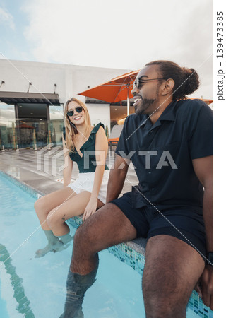 Man And Woman Bonding Casually Near Swimming Pool With Sunglasses. Two Individuals Exchanging Relaxed Conversations Beside Pool With Casual Attire And Modern City Backdrop 139473385