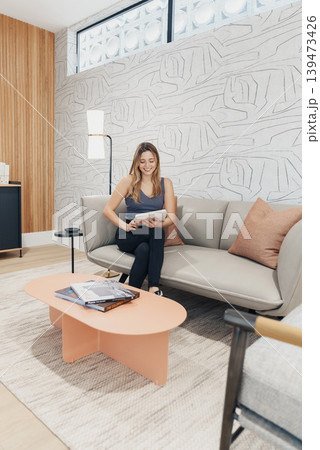 Woman On Sofa Making Phone Call, Remote Interview Candidate Participating In Video Call, Professional Posture, Resume On Table, Modern Lounge Setting With Textured Wallpaper And Soft 139473426