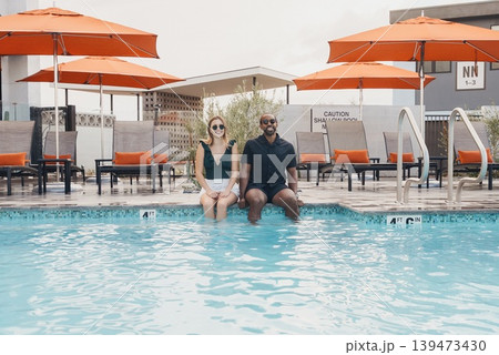 Two Friends Sitting Pool Edge Together, Bright Umbrellas And Resort Chairs Framing Clear Water Reflection, Urban Apartment Backdrop, Weekend Leisure And Smiling Candid Portrait, Modern Two Friends Sitting Pool Edge Together, Bright Umbrellas And Resort Chairs Framing Clear Water Reflection, Urban Apartment Backdrop, Weekend Leisure And Smiling Candid Portrait, Modern 139473430