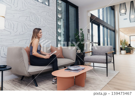 White Woman Entrepreneur Sitting On Sofa Reviewing Tablet In Bright Modern Coworking Lounge With Floortoceiling Windows, Indoor Plants, Neutral Furnishings, Relaxed Posture, Waiting Area 139473467