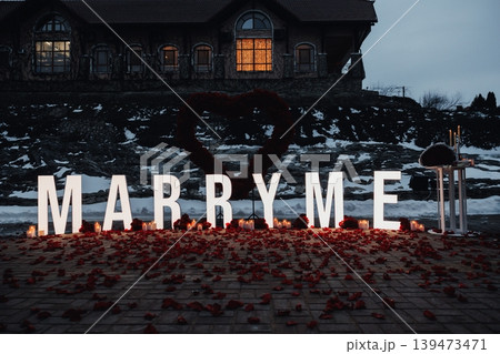 Dusk Proposal Scene With Illuminated MARRY ME Letters, Rose Petals Scattered Across Wooden Shore, Glowing Lantern, Distant House Window Casting Warm Light, Cold Evening Air, Cinematic 139473471