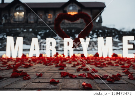 Closeup Proposal Display With Heart Wreath And Illuminated MARRY ME Letters, Rose Petals Scattered On Wooden Planks, Dramatic Glow And Vibrant Red Tones, Handcrafted Installation Staged 139473473