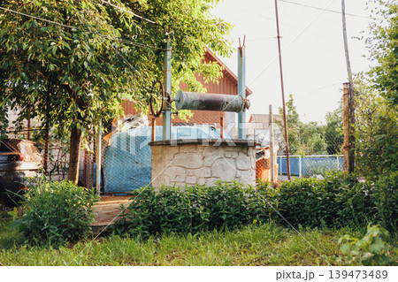 Rustic Stone Well Among Green Shrubs, Weathered Wellhead With Tiled Roof And Nearby Blue Water Tank, Overgrown Grass And Metal Pipework With Aged Fence Posts, Nostalgic Rural Atmosphere 139473489