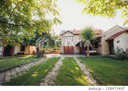 Spacious Stone Courtyard Framed By Trees And Trimmed Lawn, Dual Grass Tracks Leading To Wooden Garage Doors, Calm Midday Light, Vintage Charm And Tidy Landscape Suited For Real Estate Spacious Stone Courtyard Framed By Trees And Trimmed Lawn, Dual Grass Tracks Leading To Wooden Garage Doors, Calm Midday Light, Vintage Charm And Tidy Landscape Suited For Real Estate 139473497