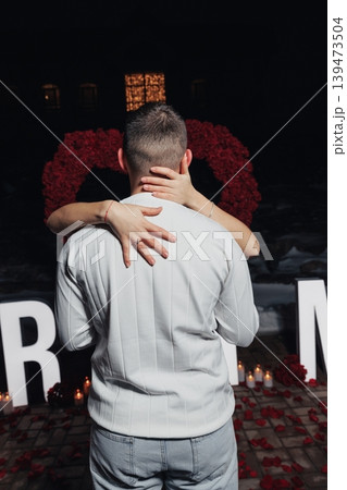 Couple Embracing In Candlelit Rose Heart Setting, Man Holding Partner Close From Behind, Hands Gripping Back, Marquee Letters Glowing, Candles And Rose Petals Scattered On Floor, Intimate 139473504