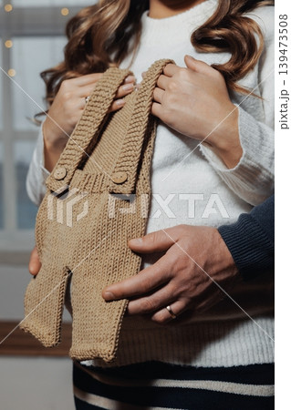 Knitted Baby Romper Held Over Belly, Closeup Of Expectant Couple Hands, Neutral Knit Fabric, Soft Natural Light, Warm Earthy Tones, Tender Touch Suggesting Anticipation And Nursery Knitted Baby Romper Held Over Belly, Closeup Of Expectant Couple Hands, Neutral Knit Fabric, Soft Natural Light, Warm Earthy Tones, Tender Touch Suggesting Anticipation And Nursery 139473508