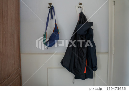 Wall hooks with hanging jacket, blue shoulder bag on white interior wall in home or hotel setting. Everyday organization, clothing storage, casual lifestyle, interior detail, accommodation concept Wall hooks with hanging jacket, blue shoulder bag on white interior wall in home or hotel setting. Everyday organization, clothing storage, casual lifestyle, interior detail, accommodation concept 139473686
