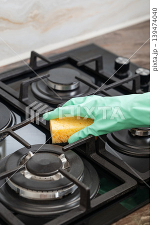 A close-up of a person's hand in a rubber glove cleaning a gas cooktop with a sponge. A close-up of a person's hand in a rubber glove cleaning a gas cooktop with a sponge. 139474040