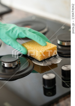 A close-up of a person's hand in a rubber glove cleaning a gas cooktop with a sponge. A close-up of a person's hand in a rubber glove cleaning a gas cooktop with a sponge. 139474042