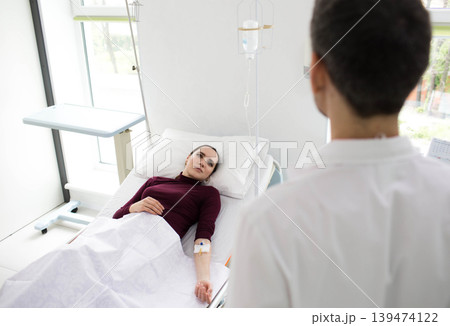 A healthcare professional attends to a patient lying in a hospital bed, equipped with an IV drip in a clean, modern clinic A healthcare professional attends to a patient lying in a hospital bed, equipped with an IV drip in a clean, modern clinic 139474122
