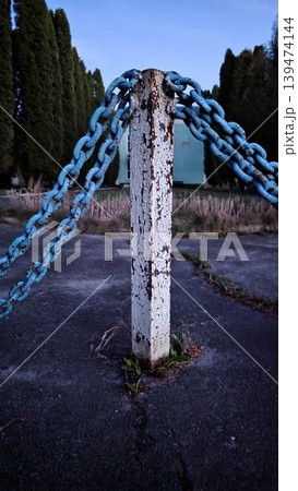 Old peeling white metal post with blue chains on cracked asphalt against row of cypress trees and turquoise building at twilight. Gloomy atmosphere of an abandoned territory Old peeling white metal post with blue chains on cracked asphalt against row of cypress trees and turquoise building at twilight. Gloomy atmosphere of an abandoned territory 139474144