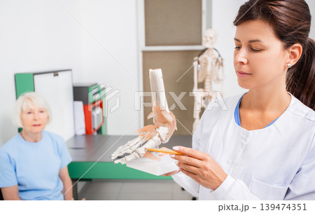 Doctor Orthopedist points to the anatomical model of the foot. Arthritis, foot injuries and pain. On background senior patient Doctor Orthopedist points to the anatomical model of the foot. Arthritis, foot injuries and pain. On background senior patient 139474351