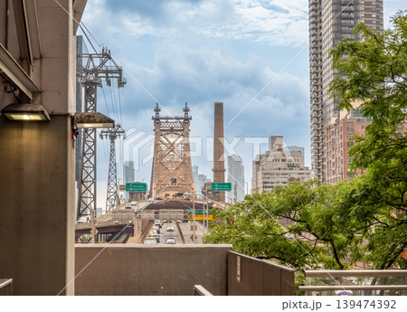 New york, usa august 17,2024. Queensboro bridge stretching across east river with traffic, roosevelt island tramway towers, and manhattan skyscrapers under a cloudy sky New york, usa august 17,2024. Queensboro bridge stretching across east river with traffic, roosevelt island tramway towers, and manhattan skyscrapers under a cloudy sky 139474392