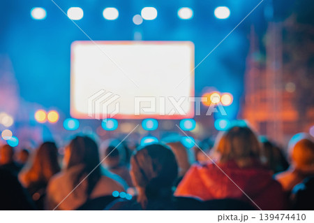 Rear view of blurred crowd watches large screen at night outdoor event with lights, bokeh, people silhouettes. Concept of screen advertising, cinema, festival, presentation, entertainment, copy space 139474410