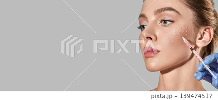 Biorevitalization procedure. Pretty woman while beauty injections with hyaluronic acid for smoothing of mimic wrinkles around eyes, over grey background with space for text 139474517