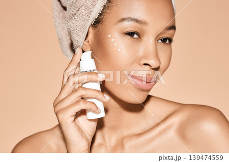 Positive curly woman applies moisturizing cream on her face, on beige background. Face cream moisturizing skin, skincare 139474559
