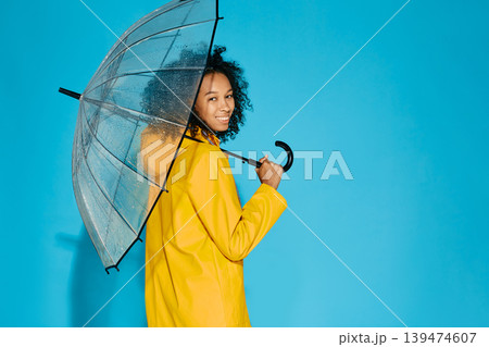 African American woman in yellow waterproof raincoat with transparent umbrella isolated on blue studio background. Autumn season concept 139474607