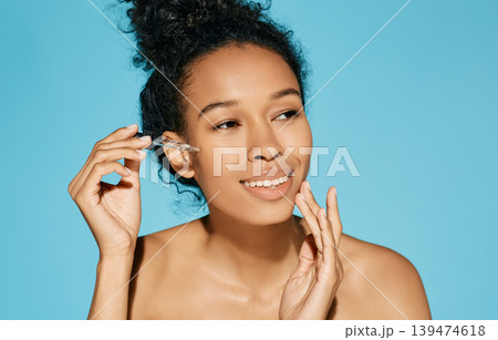Facial skin care with treatment cosmetic oil and serum. Beautiful curly African woman applying serum on her face, isolated on blue background 139474618