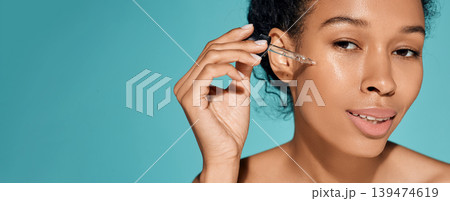 African woman holding pipette dropper with essential oil or serum near her face for hydrating and moisturizing skin 139474619