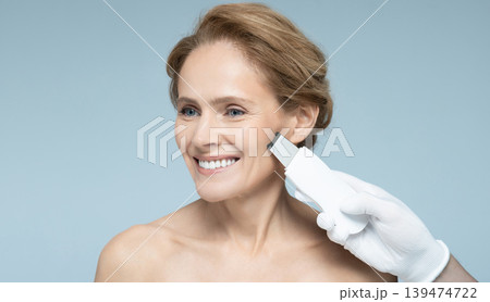 Cosmetologist using ultrasound scrubber treats skin of mature woman, removing acne and cleansing facial skin, on blue background. Ultrasonic facial peeling 139474722