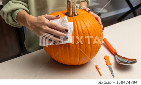 Woman cleaning a bright orange pumpkin with a cloth, surrounded by carving tools on a kitchen countertop in preparation for Halloween festivities Woman cleaning a bright orange pumpkin with a cloth, surrounded by carving tools on a kitchen countertop in preparation for Halloween festivities 139474794