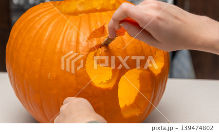 Hands carving a Halloween pumpkin with a small knife, revealing an illuminated face design, on a table with a blurred background of festive decorations 139474802