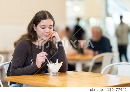 Food court girl visitor sits near table, eating ice cream in chain cafe shop Food court girl visitor sits near table, eating ice cream in chain cafe shop 139477442