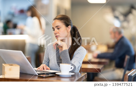 In cozy cafe hall, freelance young woman using laptop and drinks coffee. In cozy cafe hall, freelance young woman using laptop and drinks coffee. 139477521