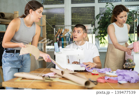 During lesson, woman teacher helps student sew, others work with fabric, blank, dress patterns 139477539