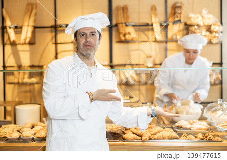 Successful baker in a white coat and chef hat invites you to buy fresh bread and buns Successful baker in a white coat and chef hat invites you to buy fresh bread and buns 139477615