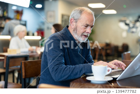 Older man is working on a laptop in a cozy cafe and drinking coffee 139477782