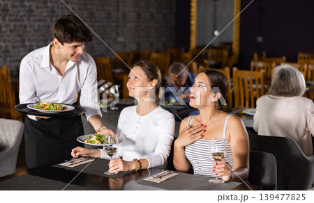 Young waiter serving ordered dishes to women in restaurant Young waiter serving ordered dishes to women in restaurant 139477825