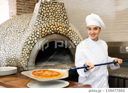 Portrait of young woman chef working in restaurant kitchen, getting ready pizza out of oven Portrait of young woman chef working in restaurant kitchen, getting ready pizza out of oven 139477860
