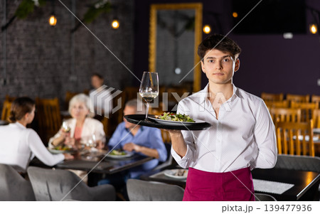 Young waiter guy is standing with order tray in spacious guest room in restaurant. Young waiter guy is standing with order tray in spacious guest room in restaurant. 139477936